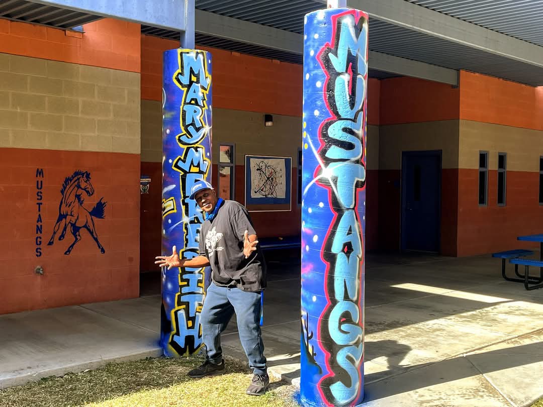 Artist stands in front of two pillars painted in a street art style featuring the word: Mary Meredith Mustangs