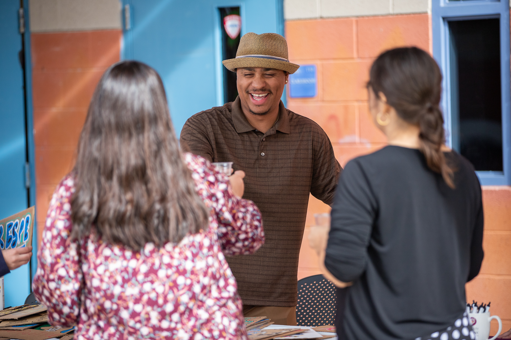 A man in a fedora smiles as he hands a woman a drink