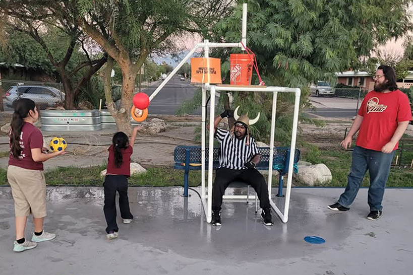 Two girls toss balls to pour water in a man in a black and white striped shirt and viking helmet
