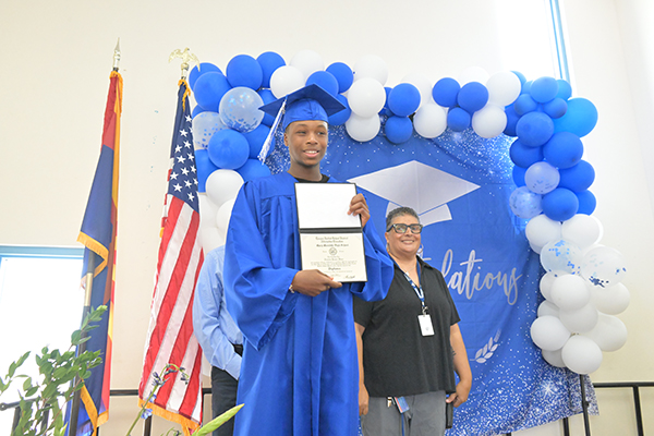 A graduate in his blue cap and gown smiles with his diploma