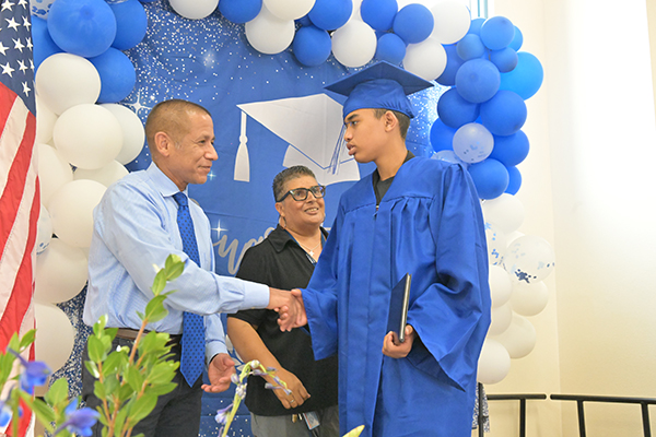 A graduate in his blue cap and gown shakes hands with the superintendent as he gets his diploma