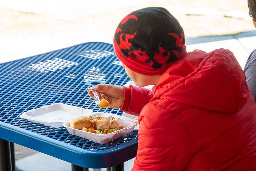 A boy in a beanie with red flames and a red jacket eats a Thanksgiving meal from a takeout box